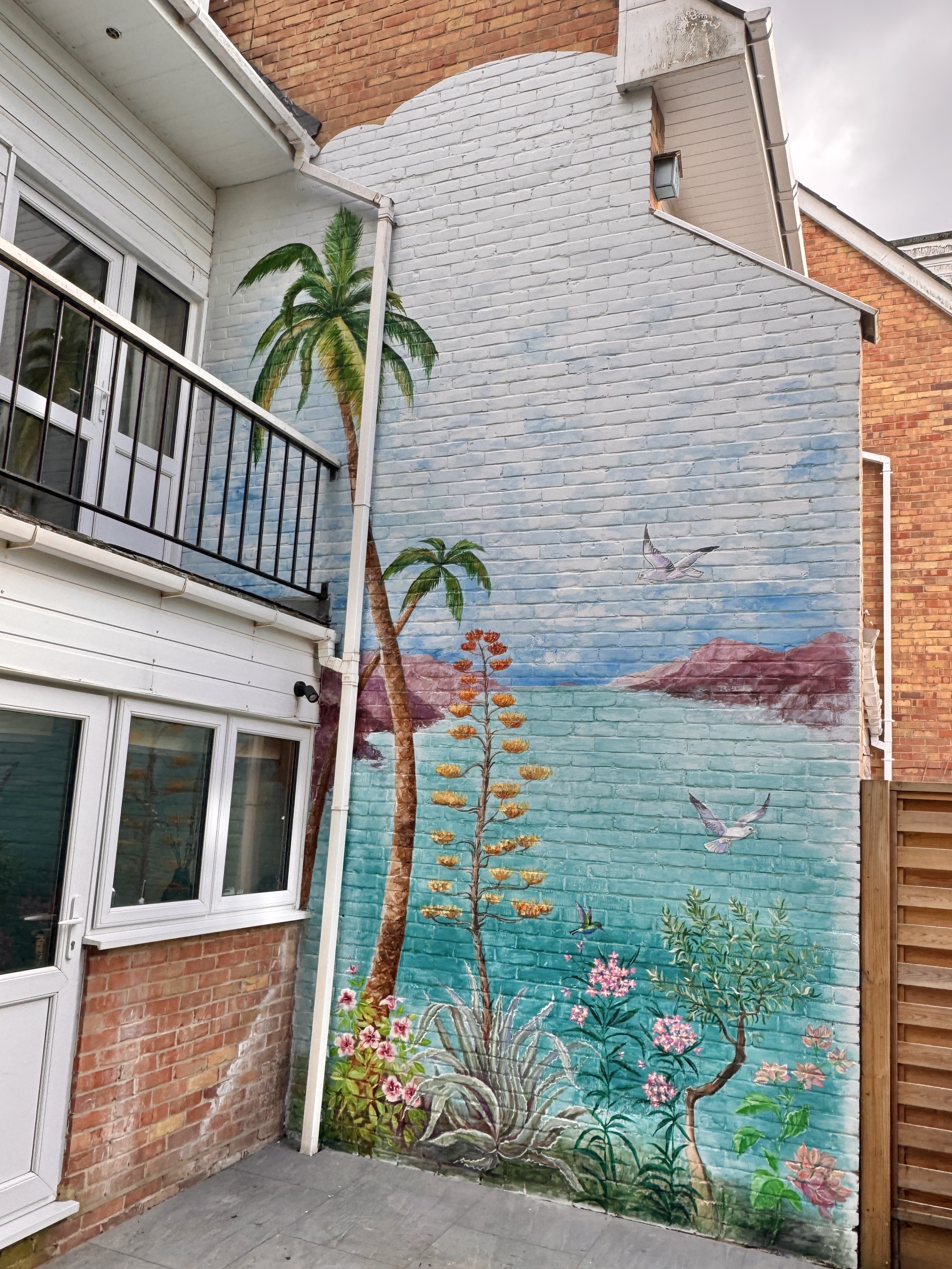 Mediterranean coastal mural with palm trees and a bay, painted on a garden patio wall in Brentwood